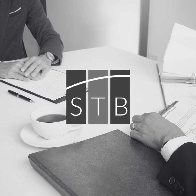 An einem Tisch sitzen 2 Personen zu einer Besprechung. In der Mitte ist das Logo von Steuerberaterin Susan Tellge-Bohne mit den Buchstaben STB abgebildet.